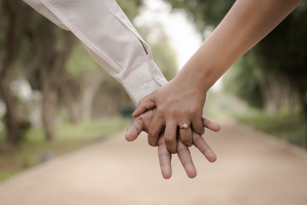 A couple holding hands along a park walkway and can see the woman’s side stone rose gold engagement ring