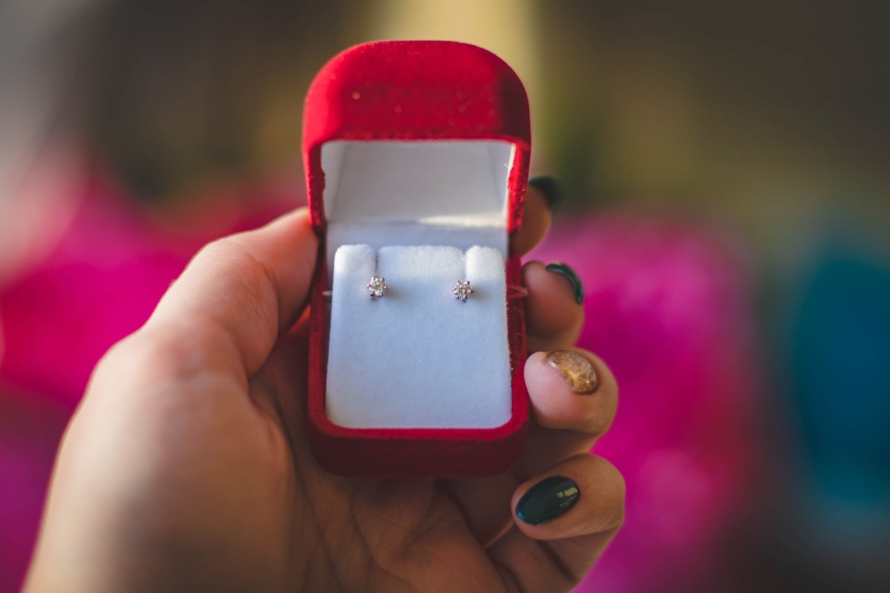 A close-up of a hand holding an open red velvet jewelry box containing a pair of small diamond stud earrings.