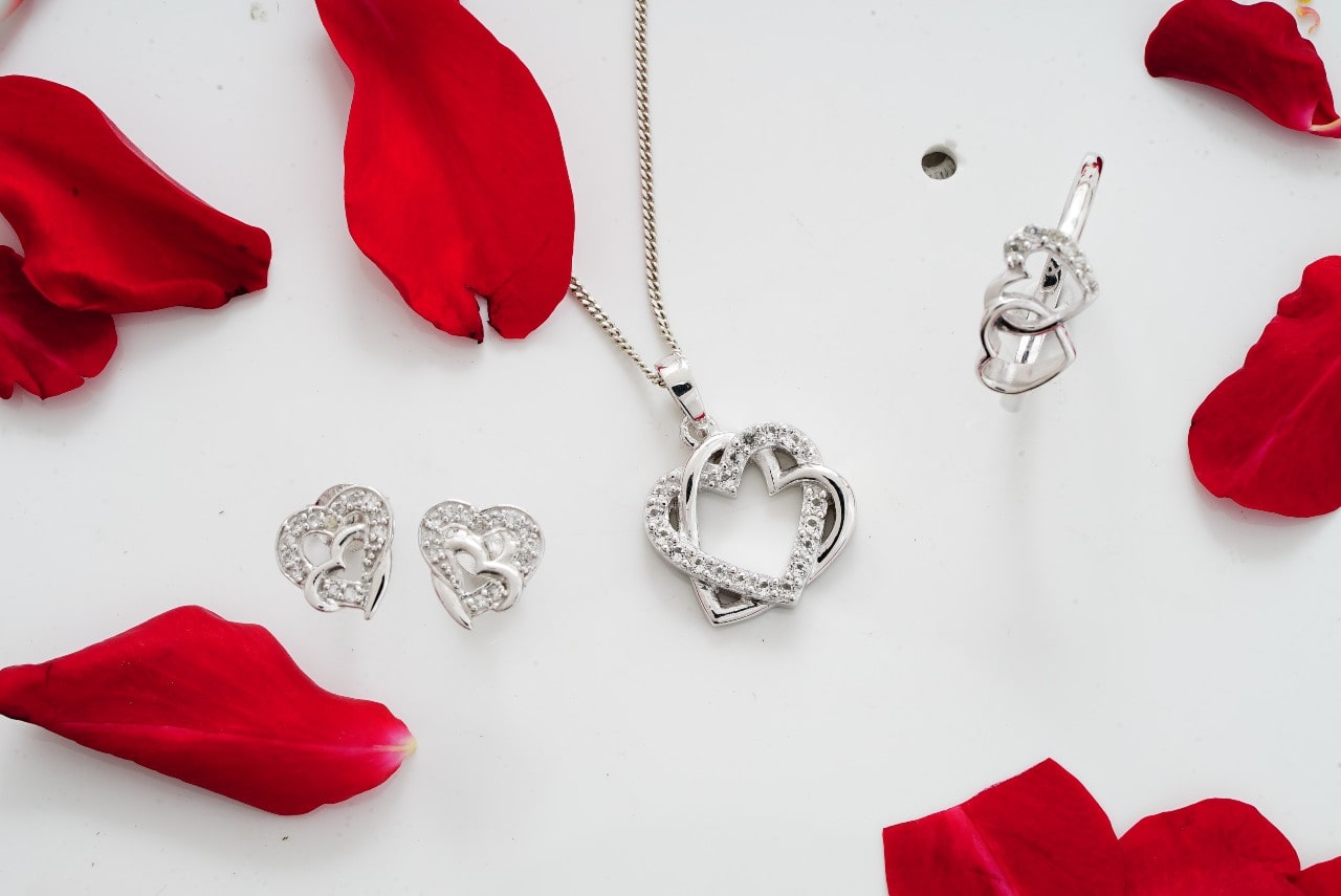 A silver heart-shaped jewelry set, including a pendant, earrings, and a ring adorned with sparkling diamonds, arranged on a white surface with scattered red rose petals